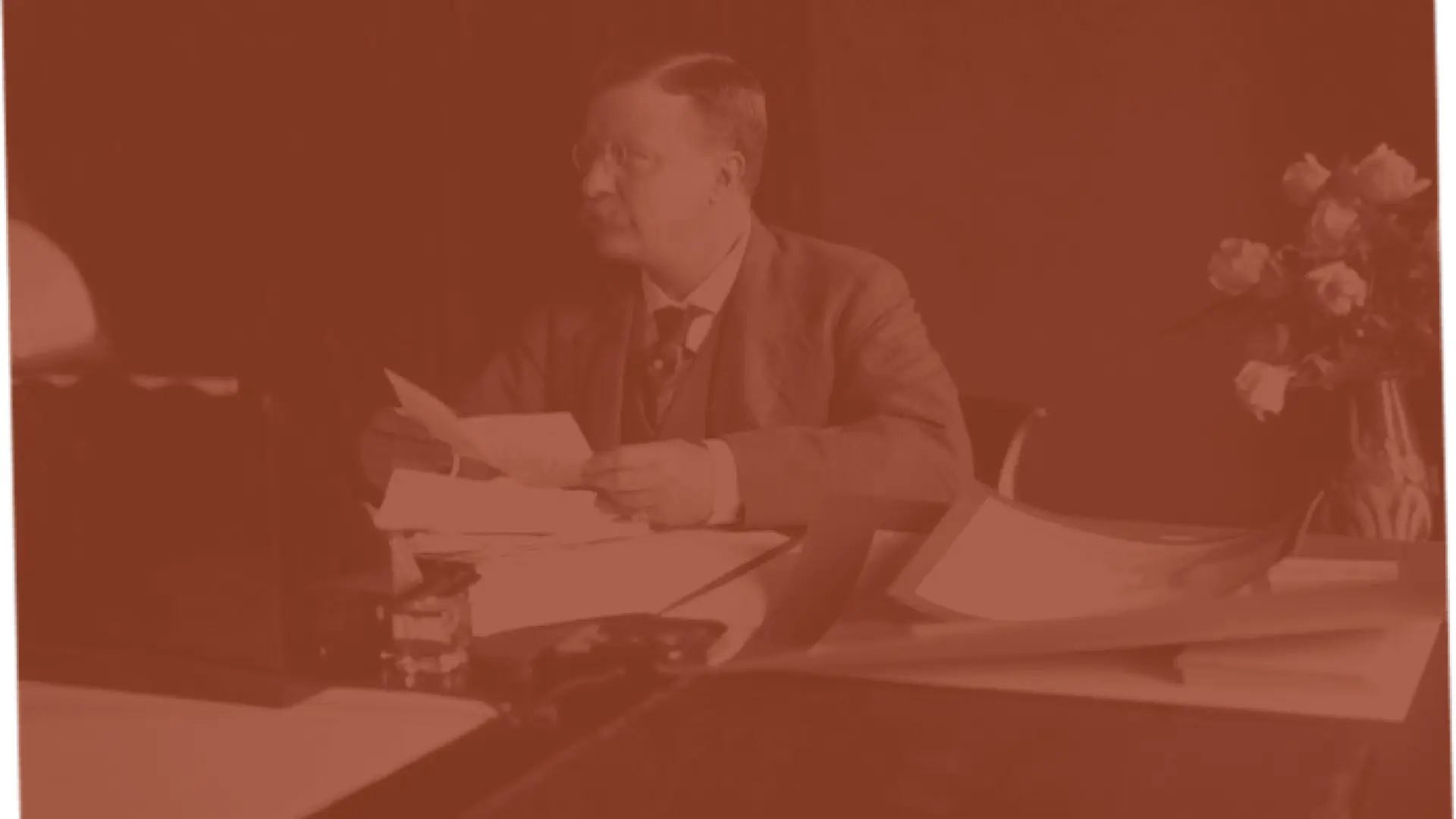 Seated man at a desk, reading papers, with flowers nearby.