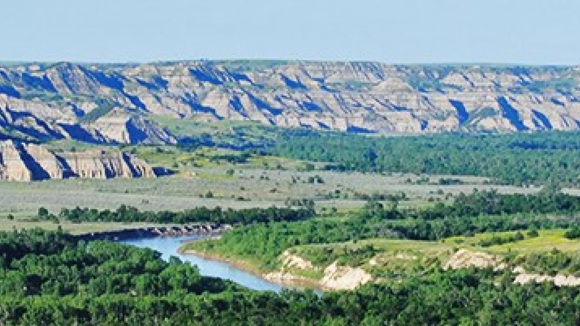 Tan and grey striped buttes rise above a green river valley in the North Unit of Theodore Roosevelt National Park