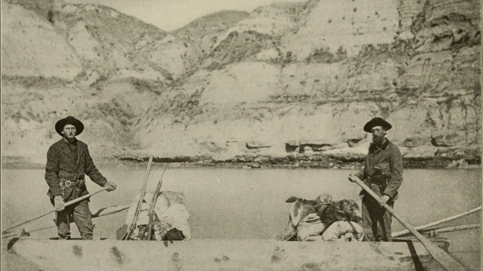 Two men poling a flat-bottomed boat loaded with gear on a river below steep clay bluffs.