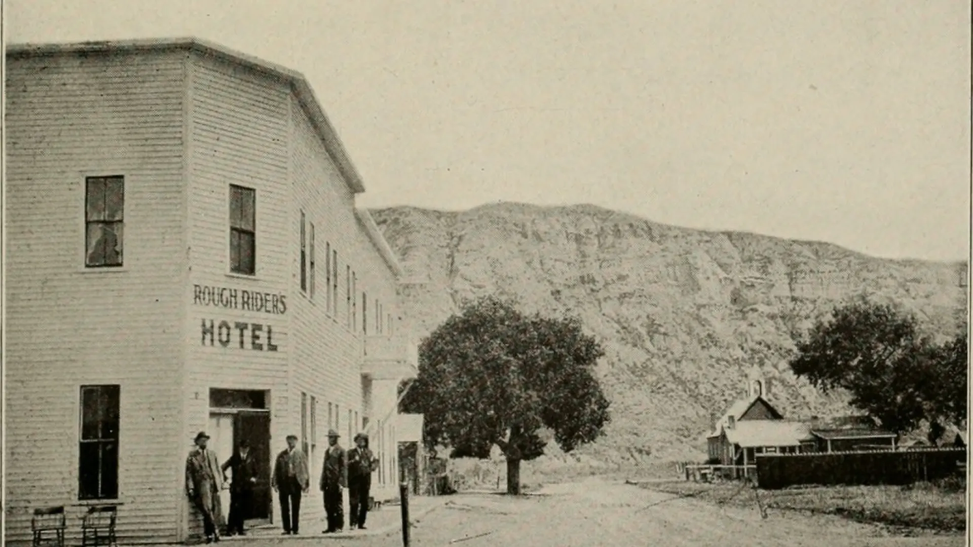 A two-story frame hotel with ROUGH RIDERS HOTEL painted on the front, men standing outside, a butte behind.