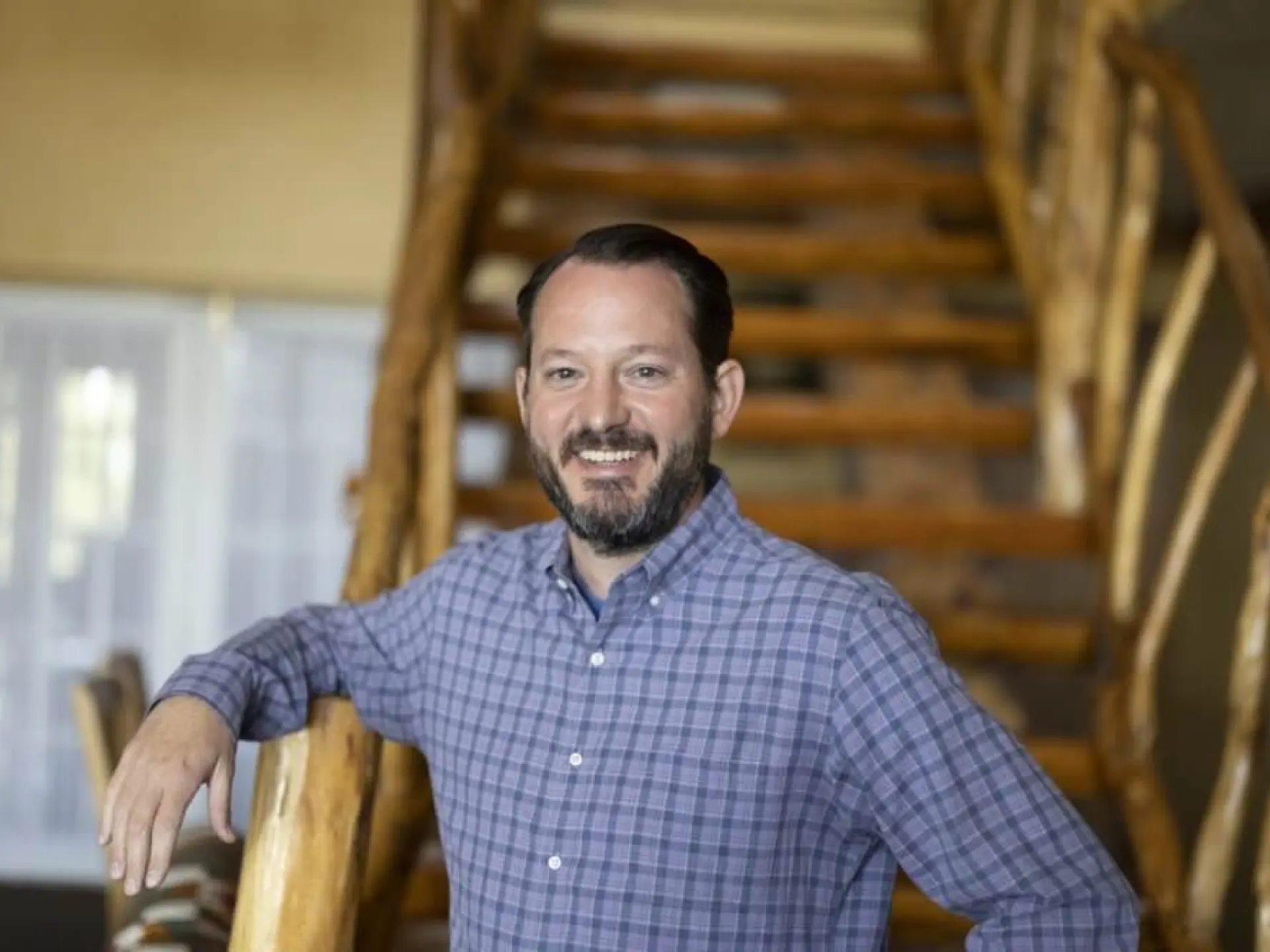 Man in a checkered shirt smiling by a wooden staircase.