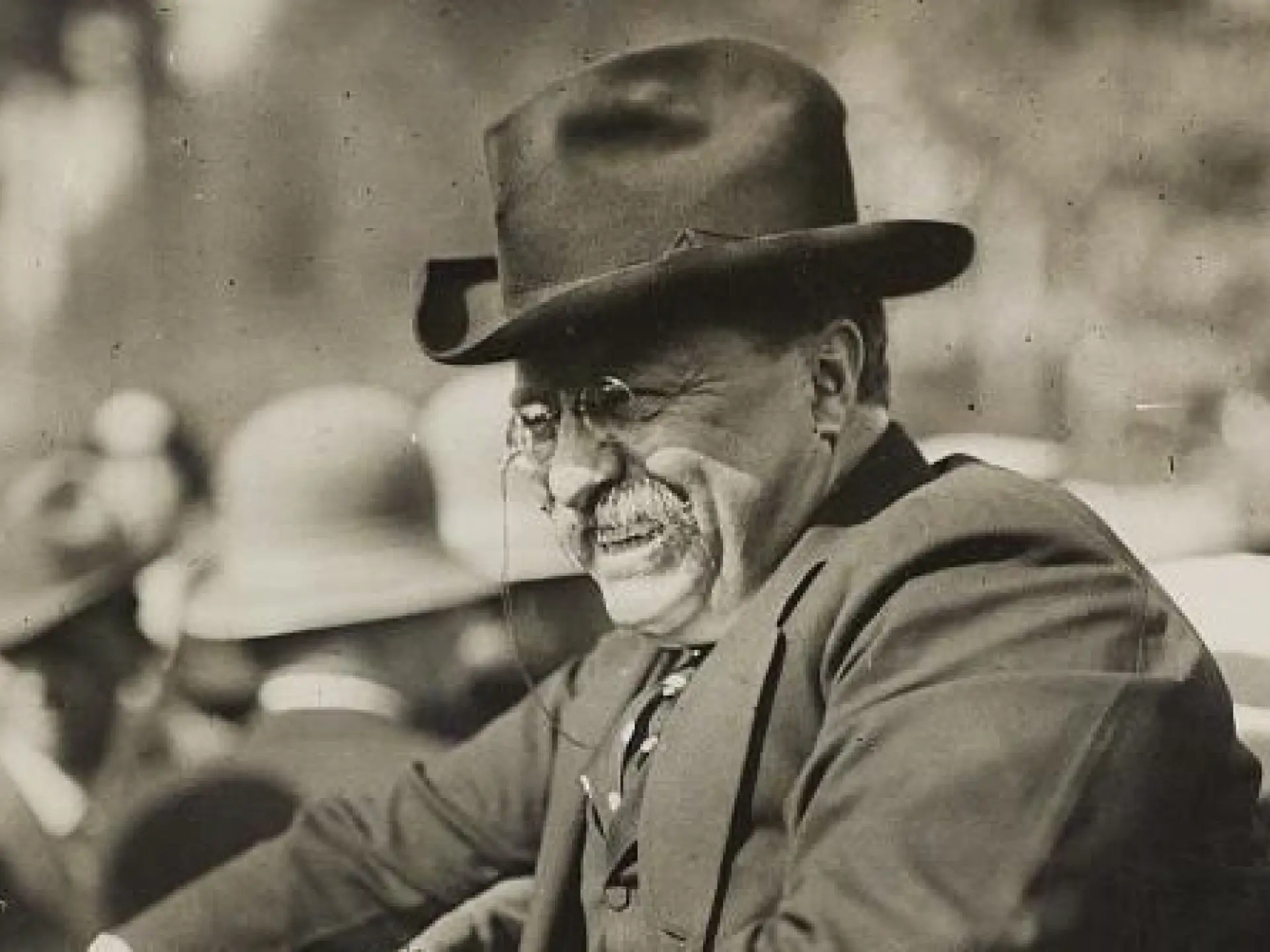 Man in a suit and hat smiling, surrounded by a crowd. Black and white photo.