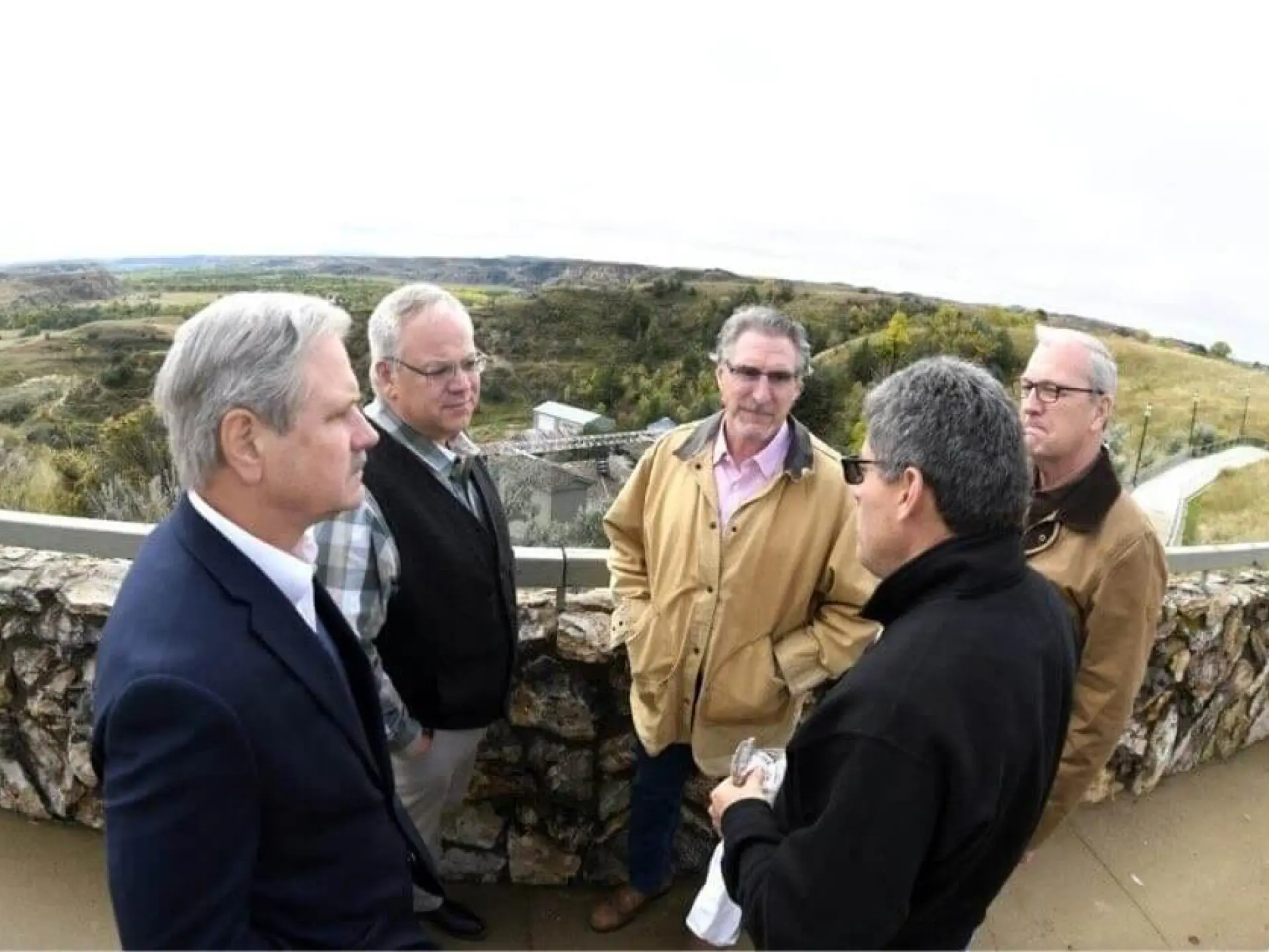 Five men in conversation on a scenic overlook with countryside views.
