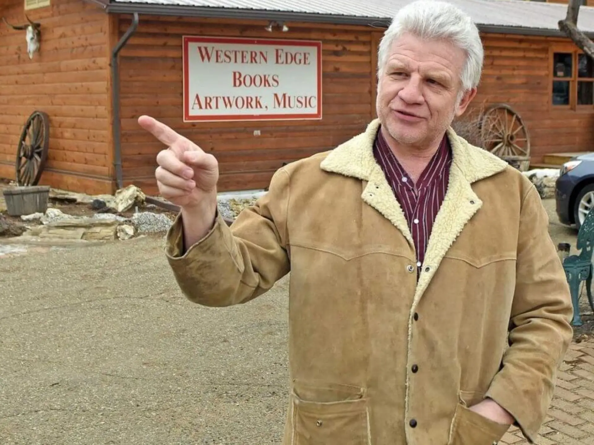 Man in a tan coat gestures outside a rustic wooden building.