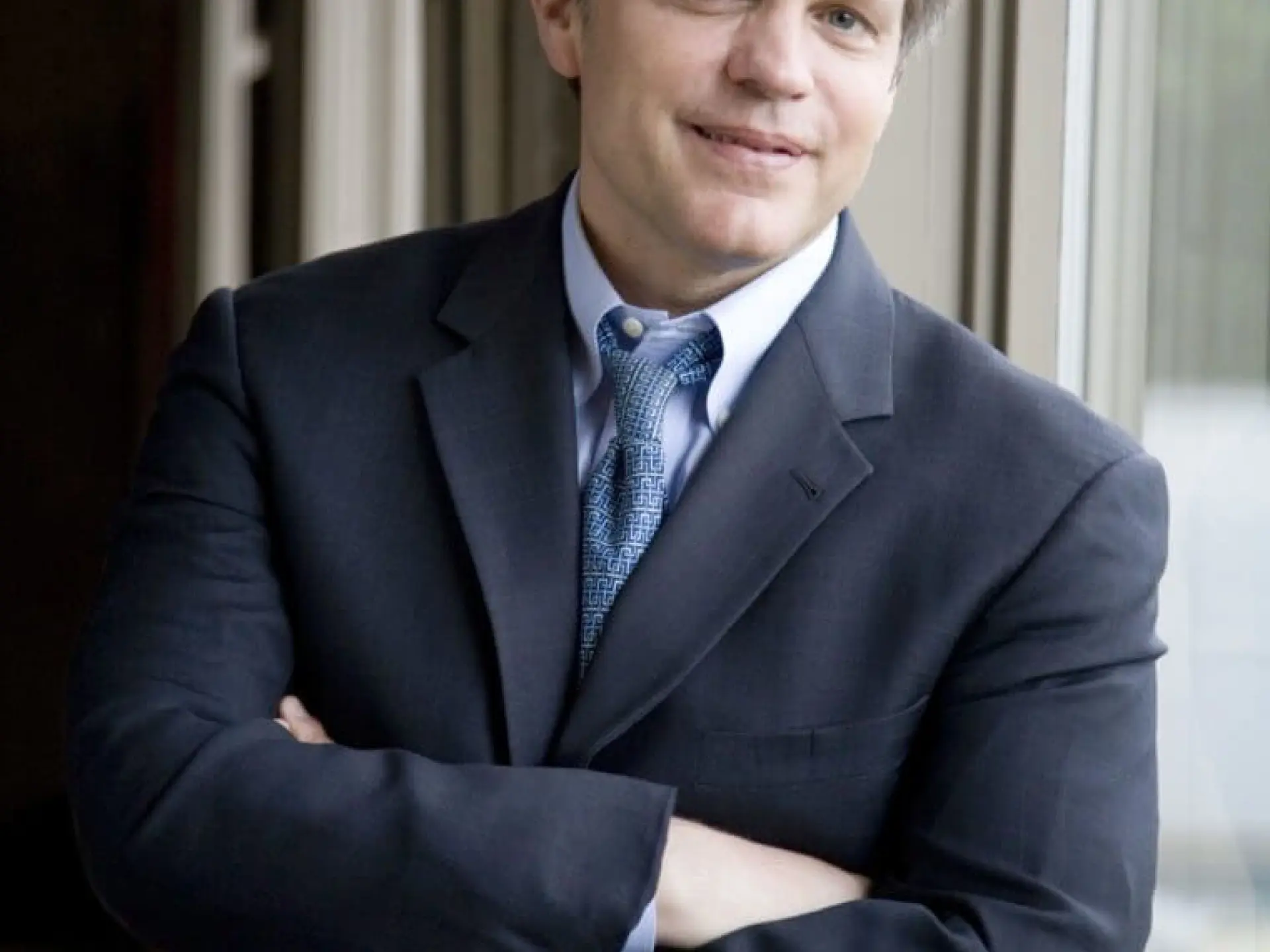 Man in a suit smiling, leaning against a window.