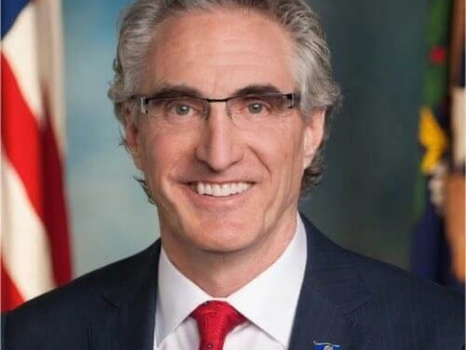 Smiling man in a suit with flags in the background.