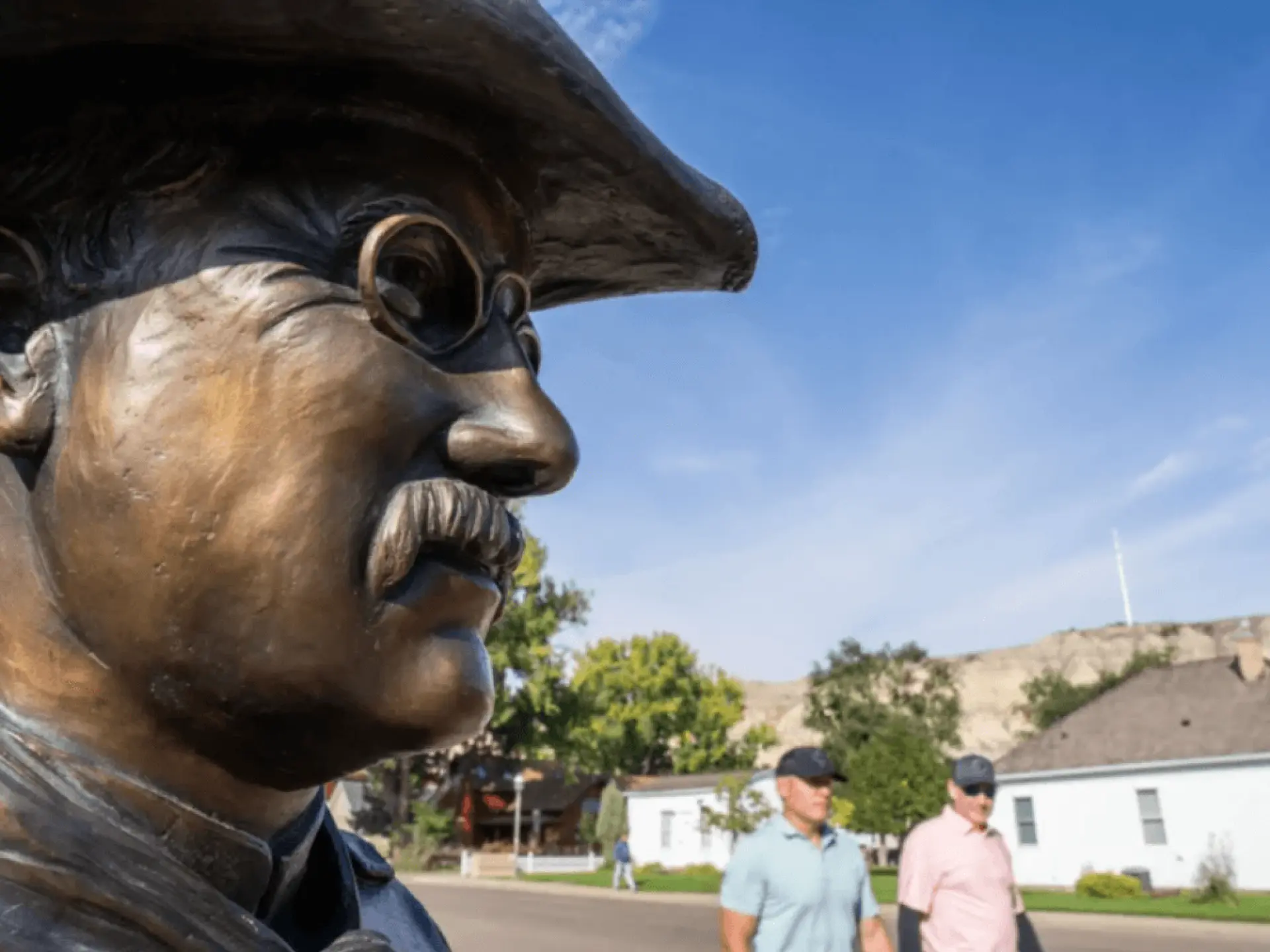 Bronze statue in profile under a clear blue sky; people walking in background.