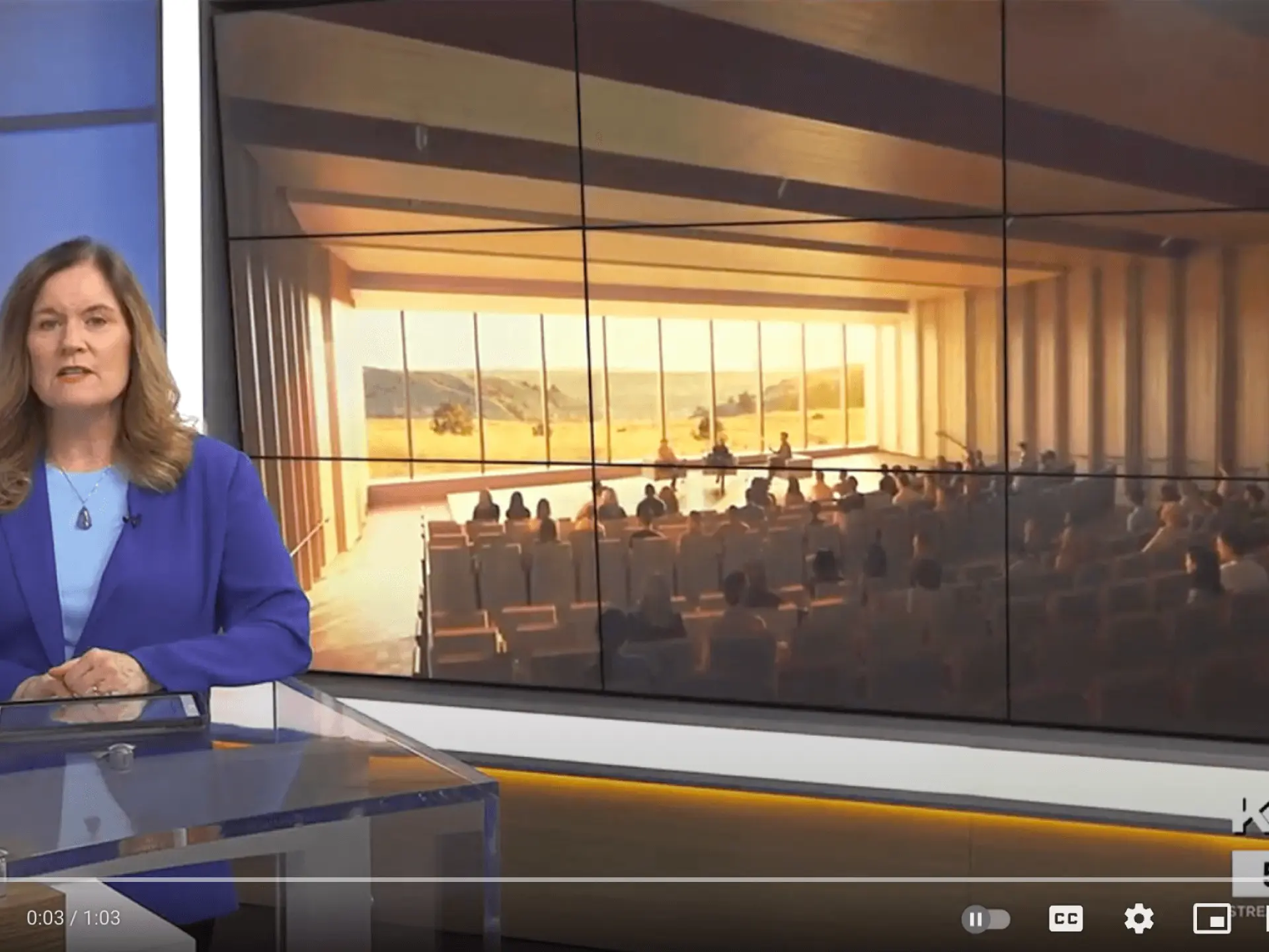News anchor in blue blazer seated at a glass desk, with auditorium image in the background.