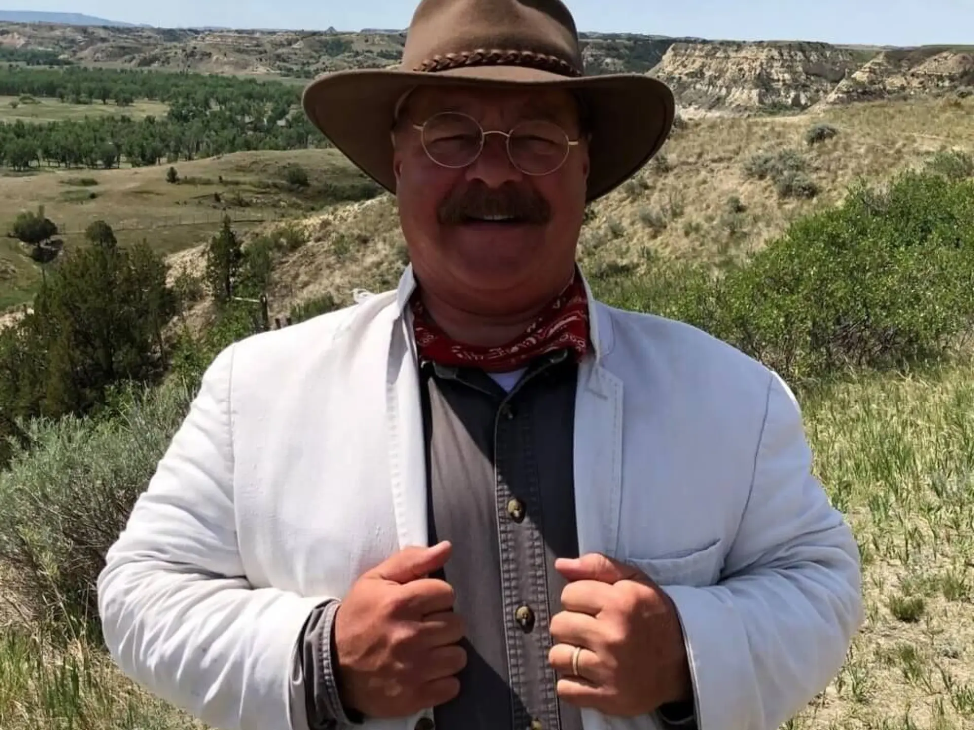 Man in cowboy hat and white jacket smiling in grassy landscape.