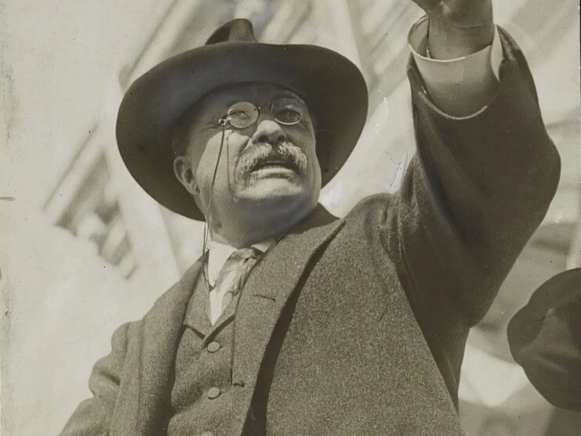 Man in suit and hat gestures upwards, standing outdoors.