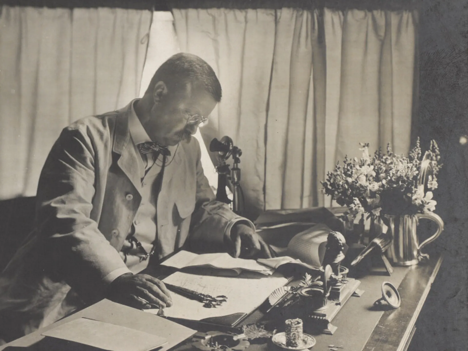 Theodore Roosevelt seated at a desk writing, with papers spread out before him
