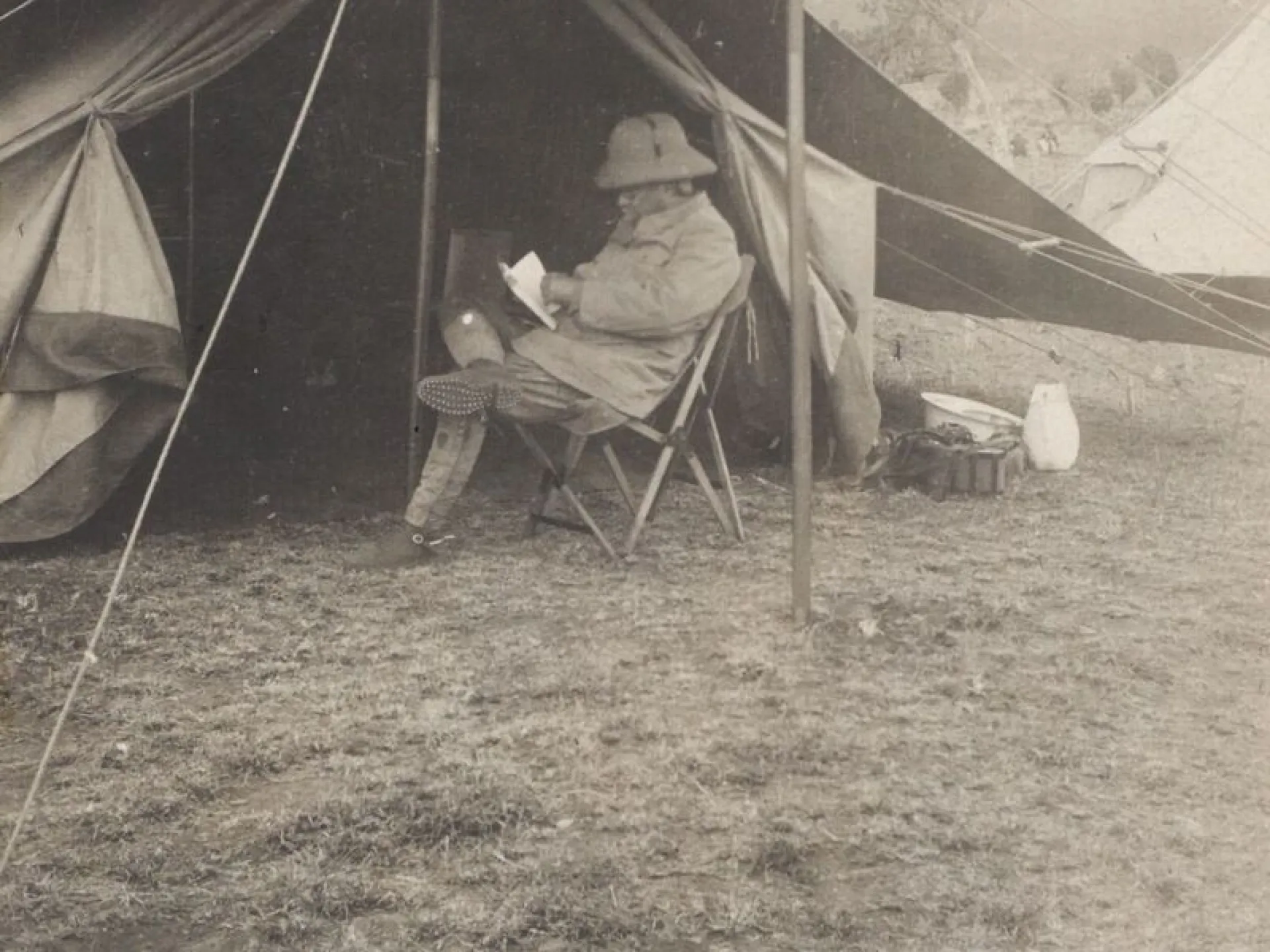 Theodore Roosevelt seated in a camp chair reading at the entrance of a tent