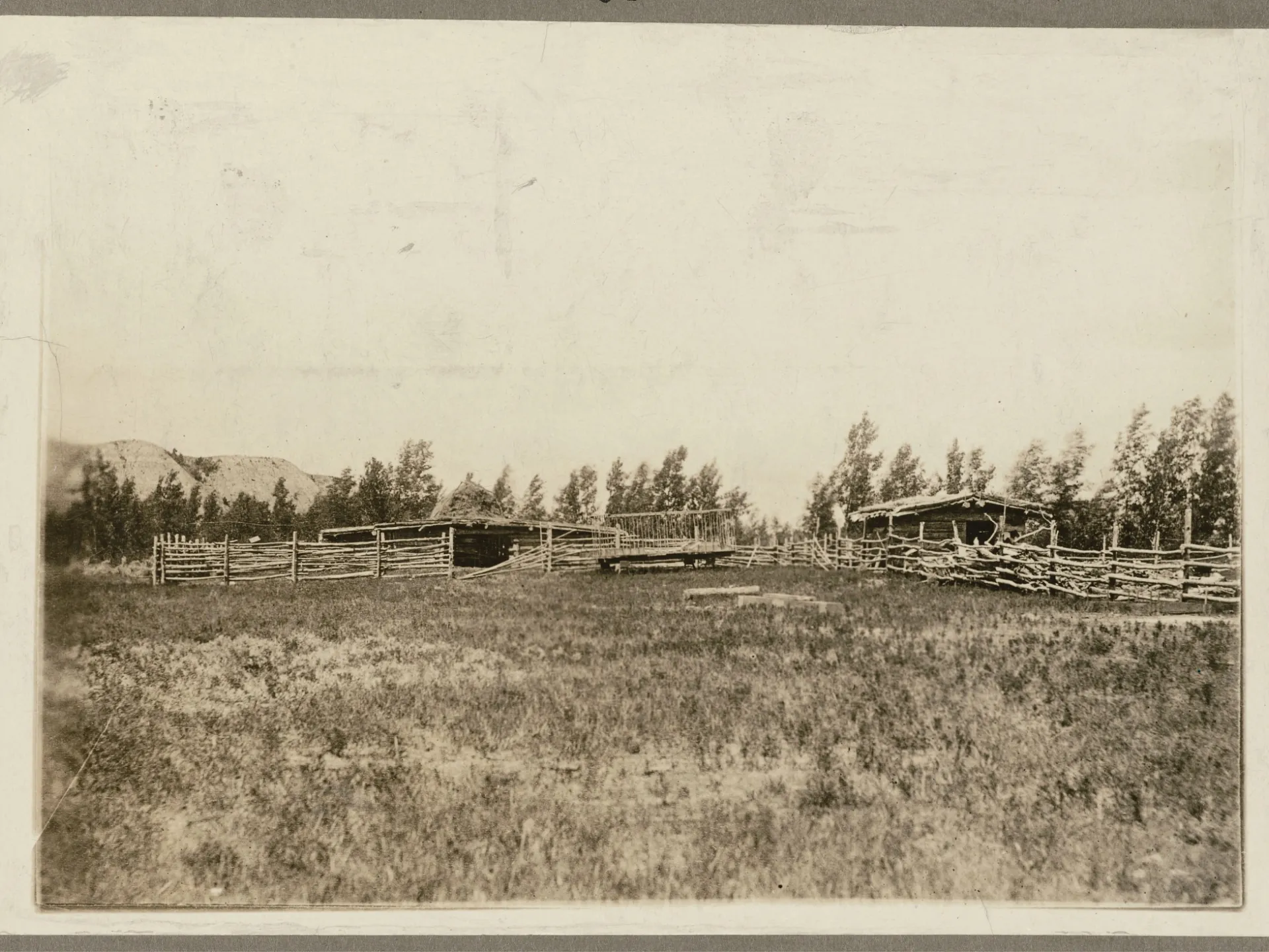 Historic photograph of the corrals and low ranch buildings at the Elkhorn Ranch, standing on open bottomland with a line of trees and bluffs behind