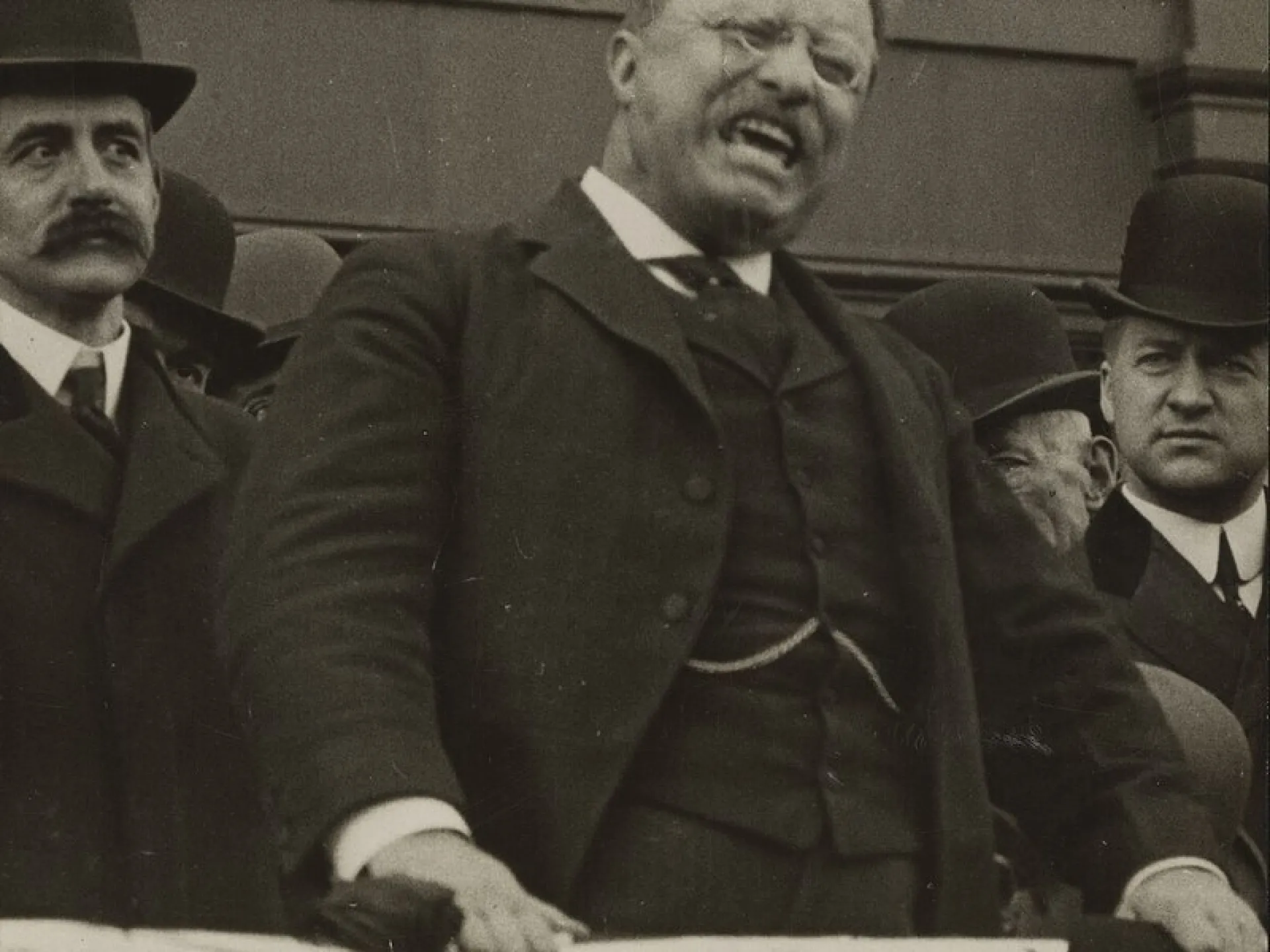 Theodore Roosevelt speaking forcefully from a platform, flanked by other men