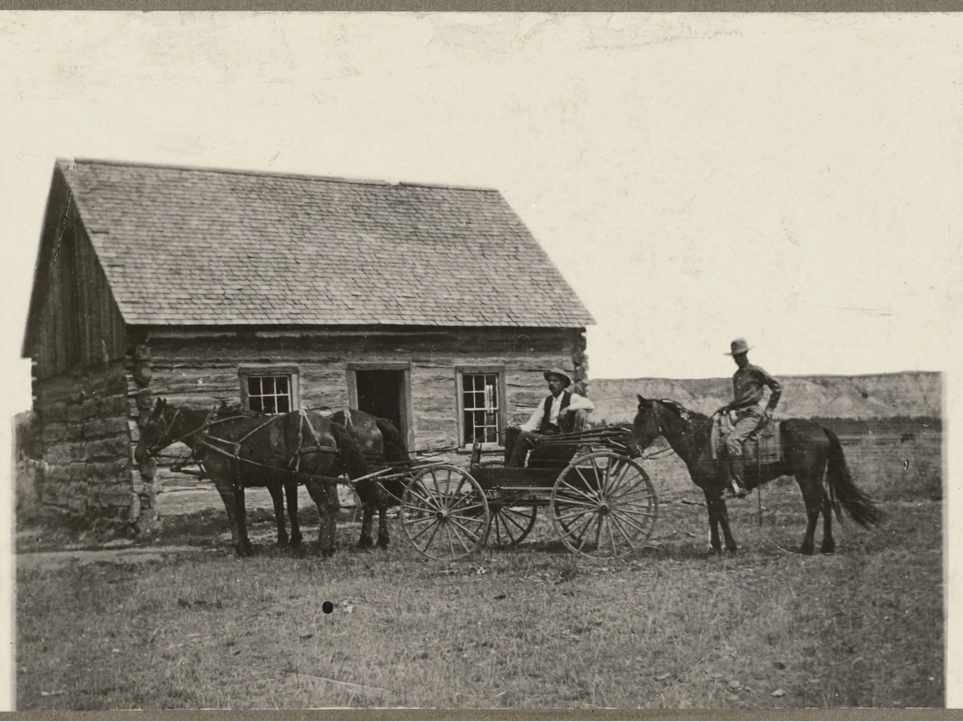 Historic photograph of the Maltese Cross Ranch cabin, a low log house with a shingled roof, with a team of horses and two mounted riders in front