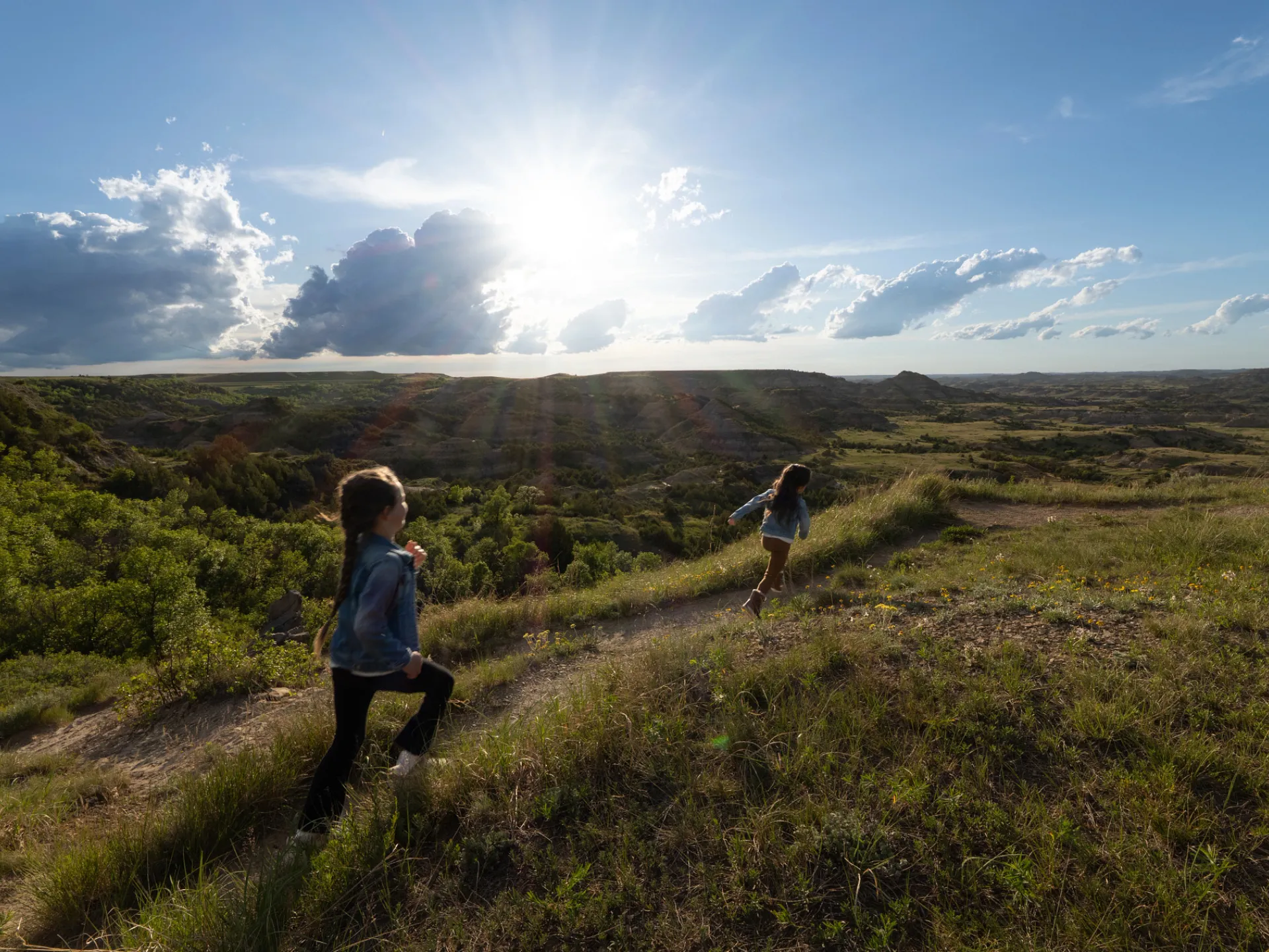 Two visitors on the trail at Painted Canyon Overlook, with layered badlands buttes stretching to the horizon under a bright sky