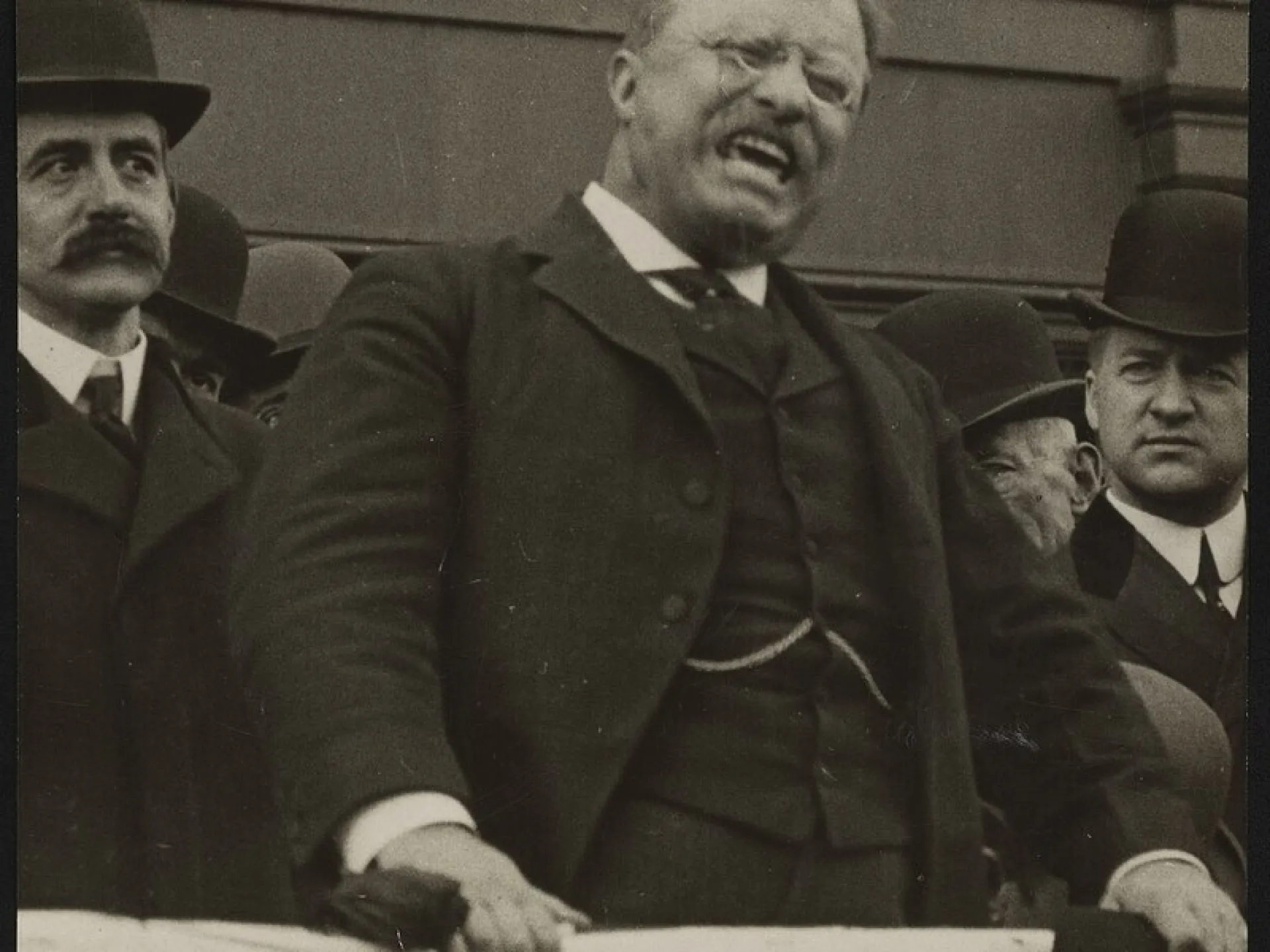 Theodore Roosevelt speaking emphatically from a platform draped with a banner