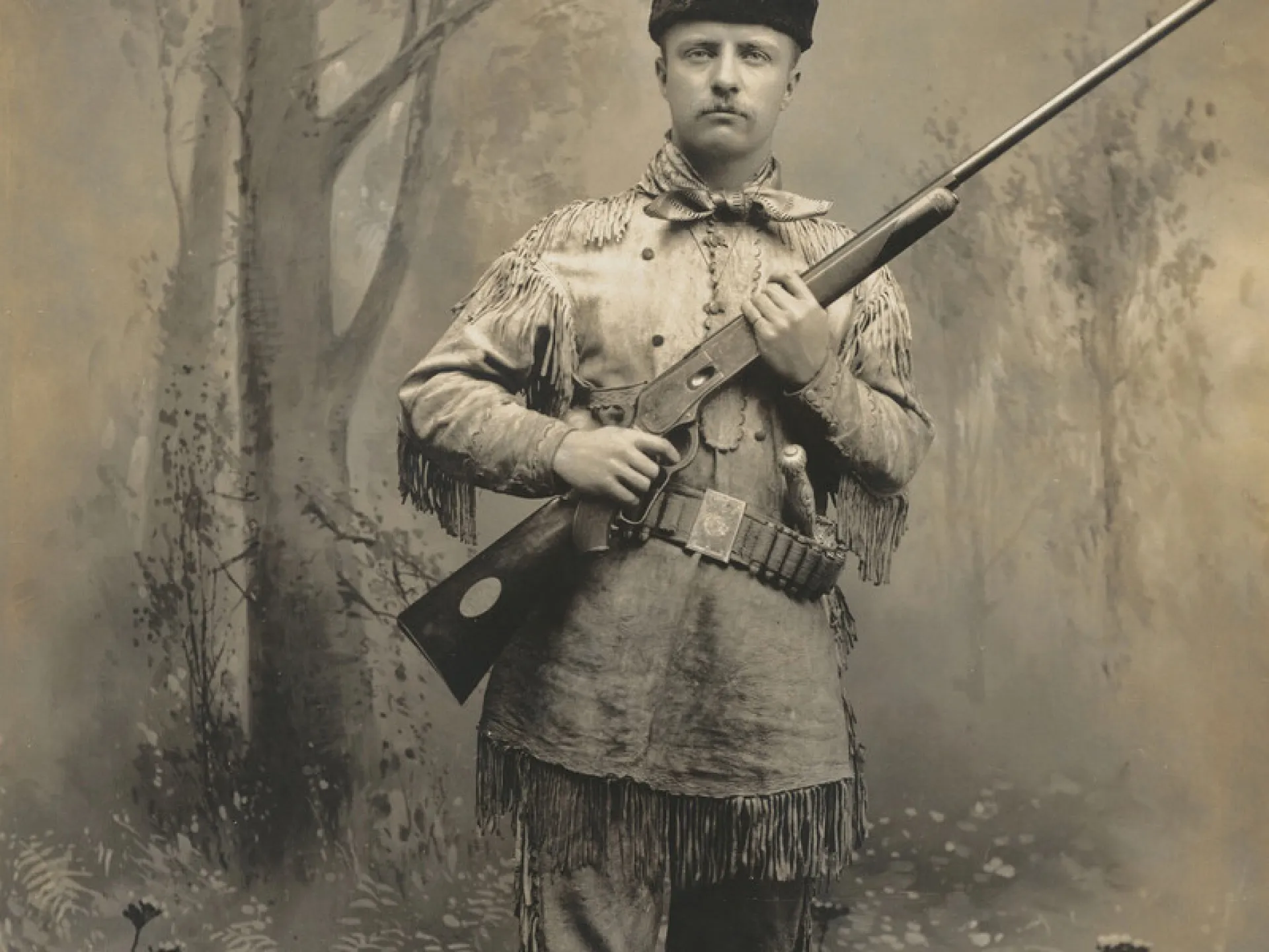Theodore Roosevelt in a fringed buckskin suit holding a rifle in a studio portrait