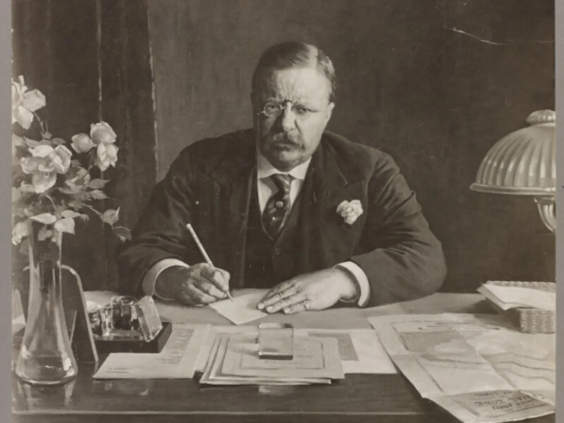 Theodore Roosevelt seated at his desk, writing among papers