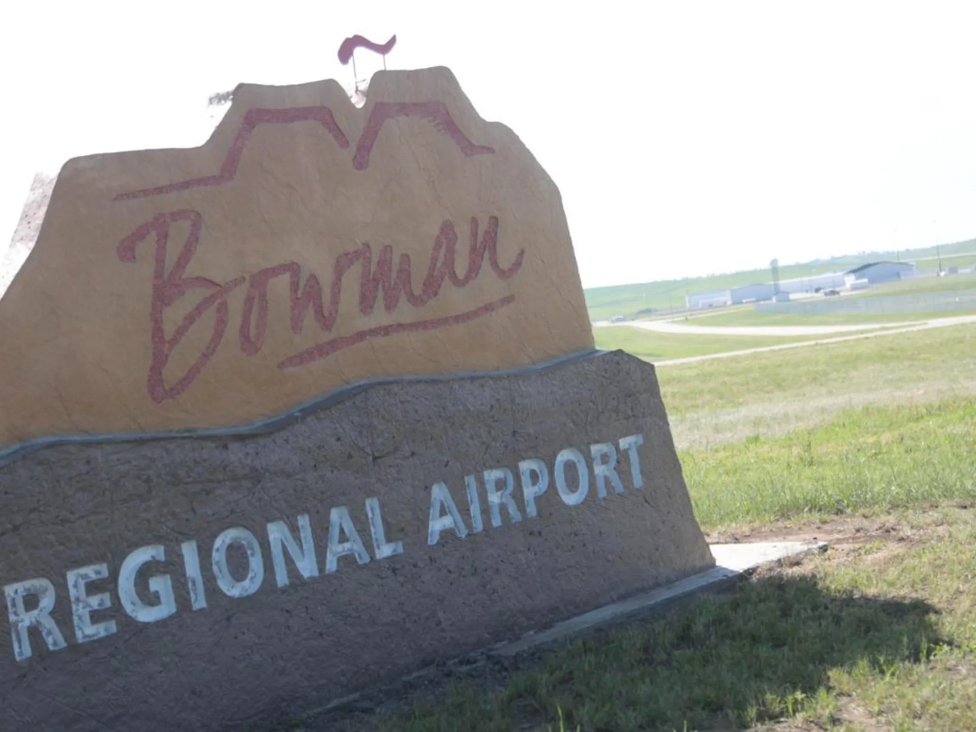 The Bowman Regional Airport sign, with the terminal and hangars on the prairie behind it