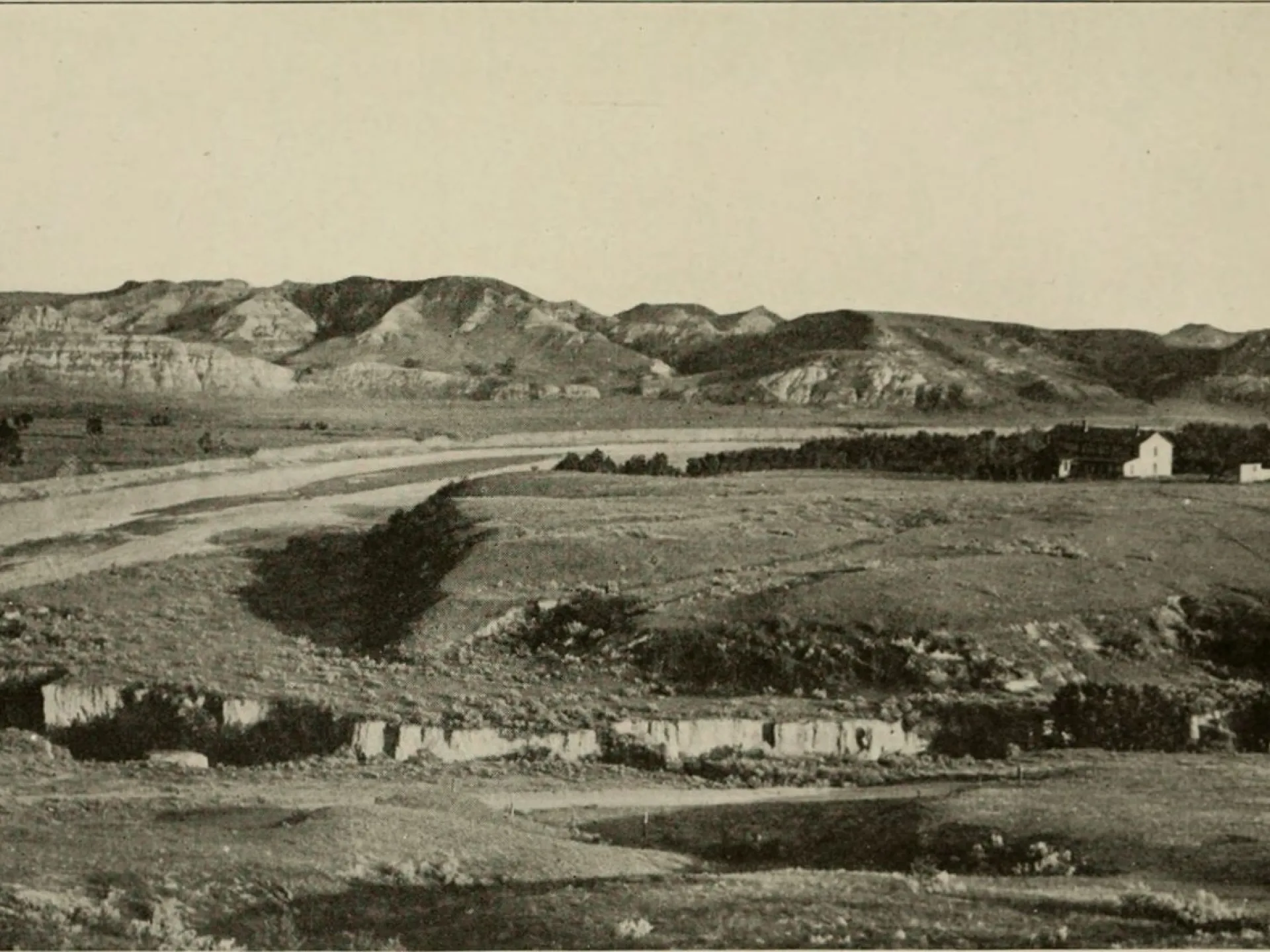 A wide river valley with eroded buttes; a large house stands on a bench above the valley floor.