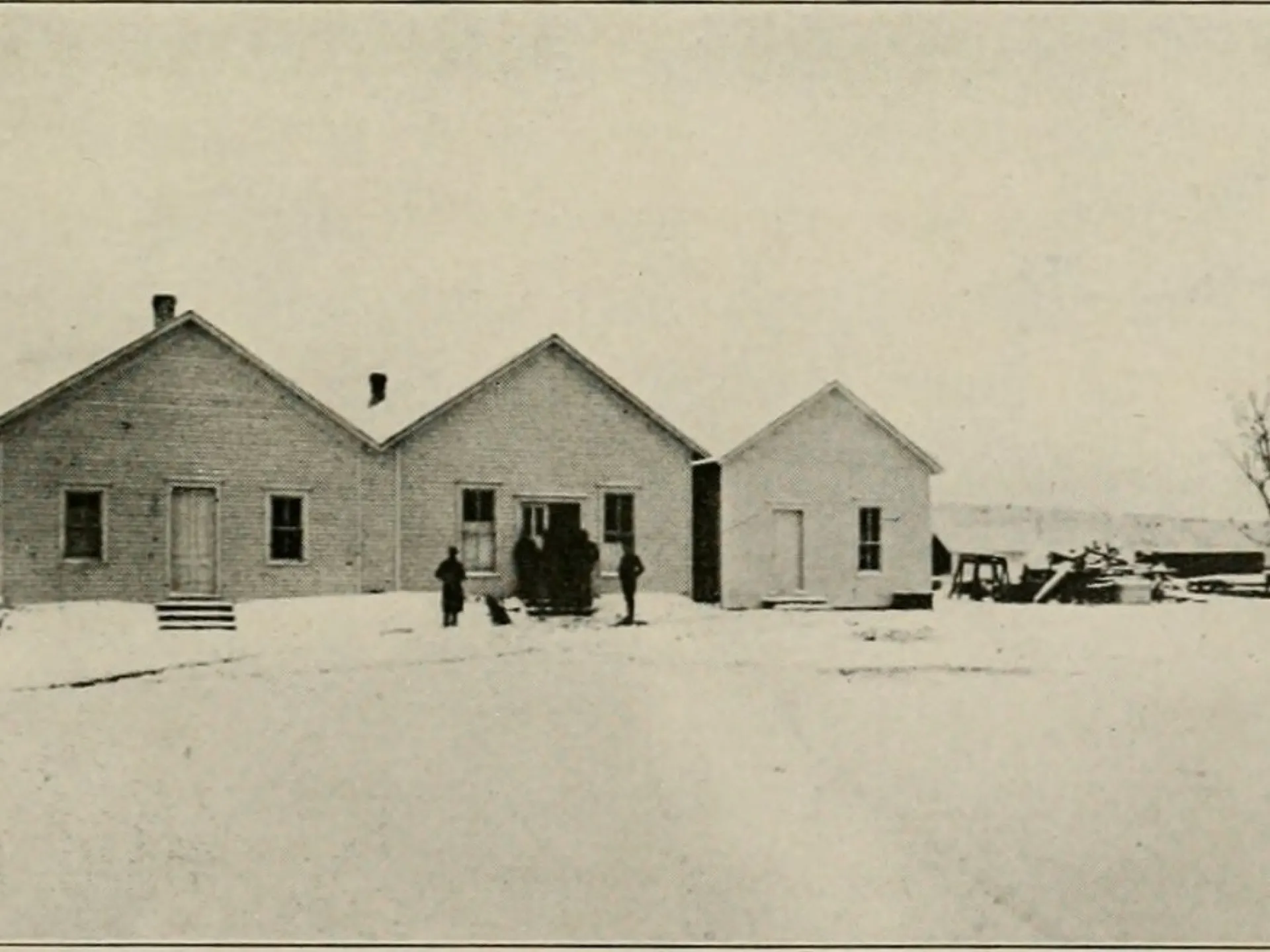 Low frame buildings in snow, a few people standing in front.
