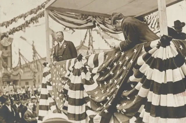 Sepia photo of men on a star-spangled podium, one speaking to a crowd.