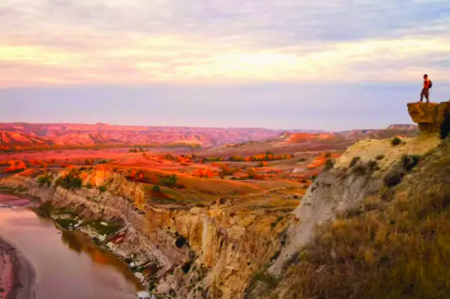 Person standing on a cliff edge at Wind Canyon in Theodore Roosevelt National Park, overlooking the Little Missouri River and colorful Badlands at sunset.