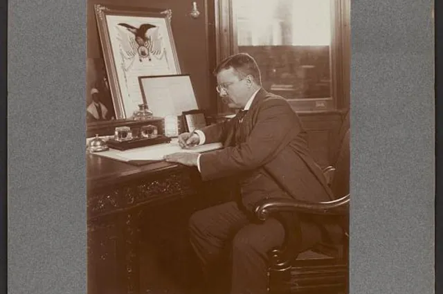 President Theodore Roosevelt seated at a desk with a pen, signing a document