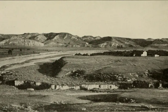 A wide river valley with eroded buttes; a large house stands on a bench above the valley floor.