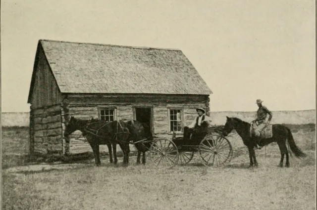 A small log cabin with a shingled roof on open prairie, a horse and buggy and a rider in front.