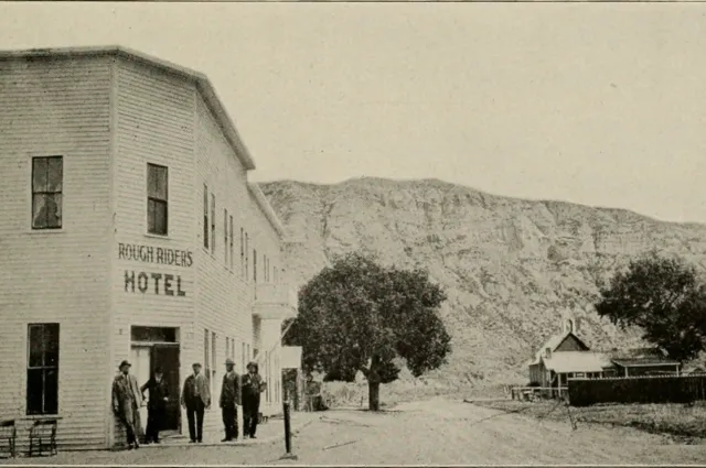 A two-story frame hotel with ROUGH RIDERS HOTEL painted on the front, men standing outside, a butte behind.