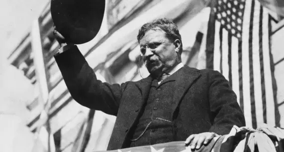 Man raising hat, speaking energetically from a balcony with American flag in the background.