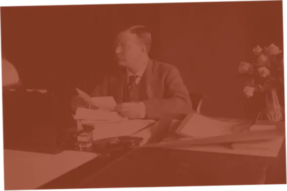 Seated man at a desk, reading papers, with flowers nearby.