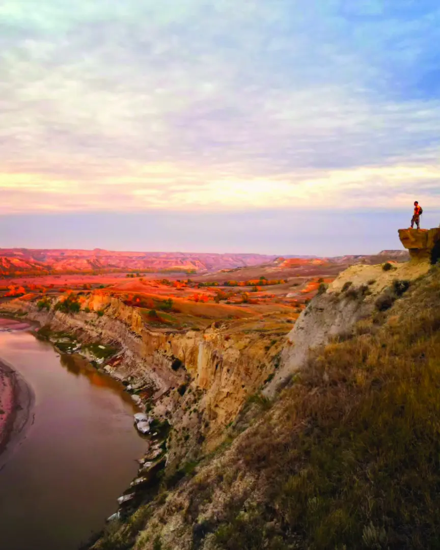 Person standing on a cliff edge at Wind Canyon in Theodore Roosevelt National Park, overlooking the Little Missouri River and colorful Badlands at sunset.