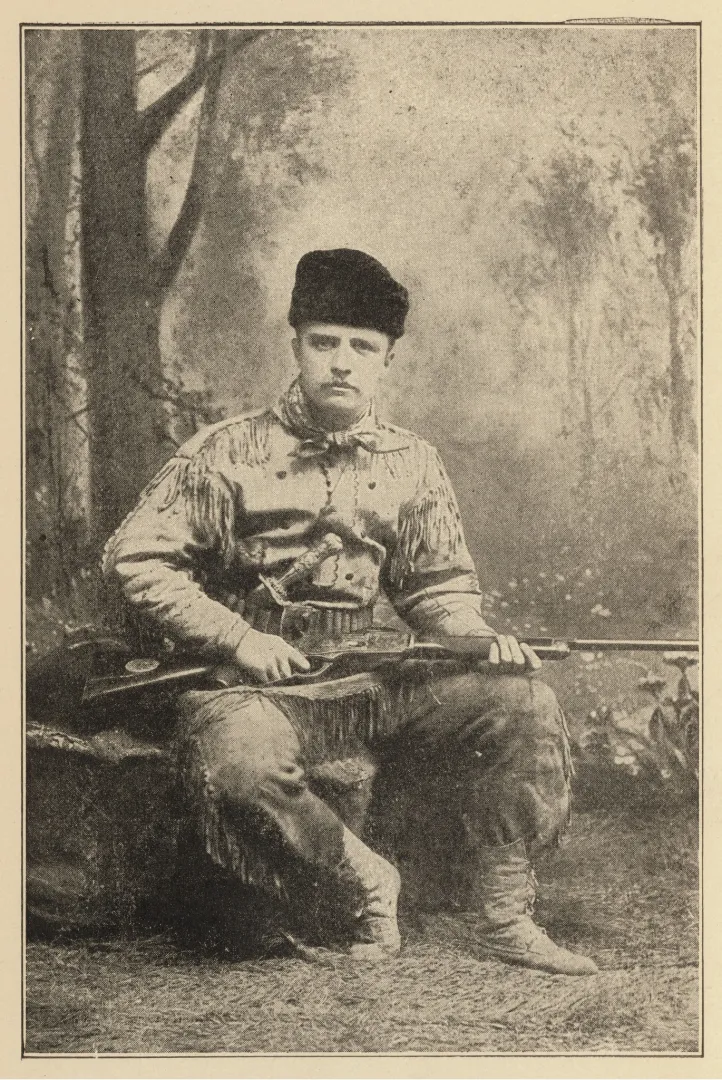 Theodore Roosevelt seated in a fringed buckskin hunting suit, holding a rifle across his lap