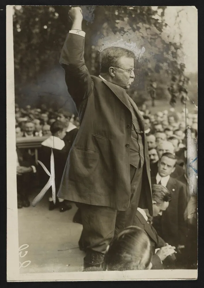 Theodore Roosevelt speaking with one arm raised to a crowd of citizens