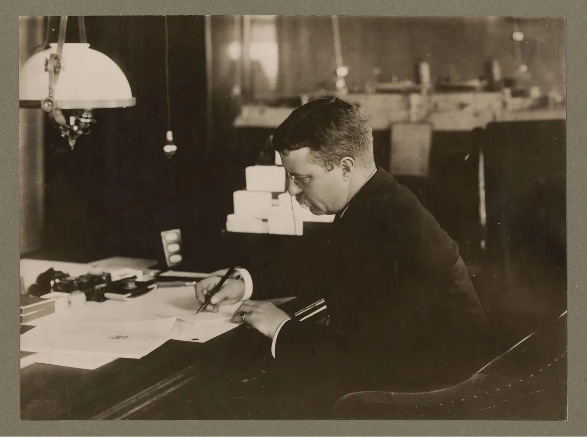 Theodore Roosevelt seated at a desk writing in a book-lined study