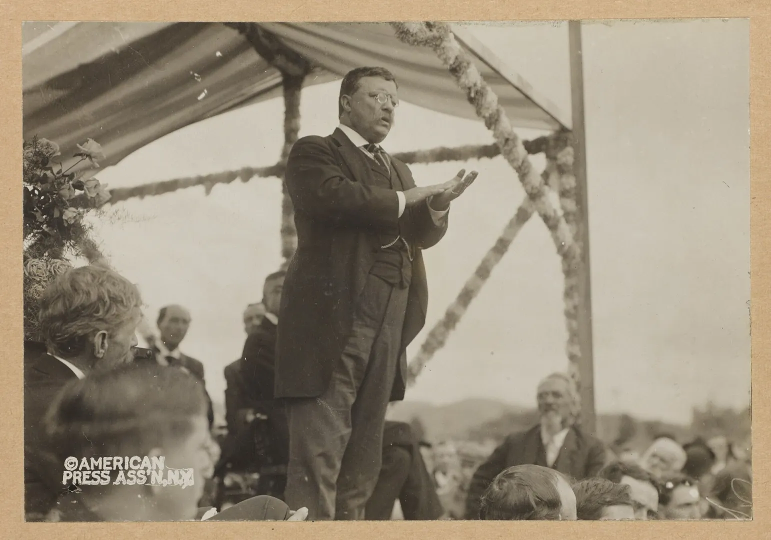 Theodore Roosevelt gesturing as he delivers a speech from a platform to a crowd