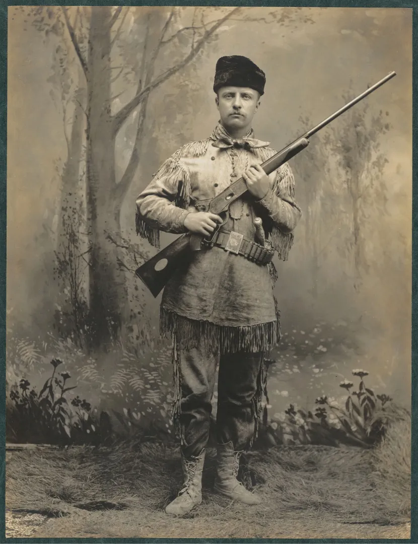 Theodore Roosevelt standing in a fringed buckskin suit holding a hunting rifle
