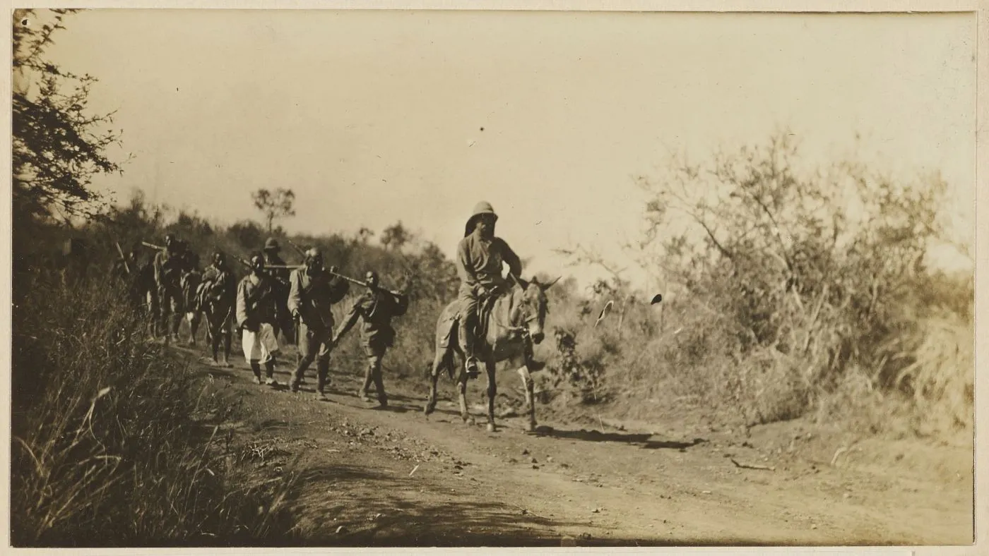 Theodore Roosevelt on horseback leading a line of safari porters along a bush trail in Uganda.