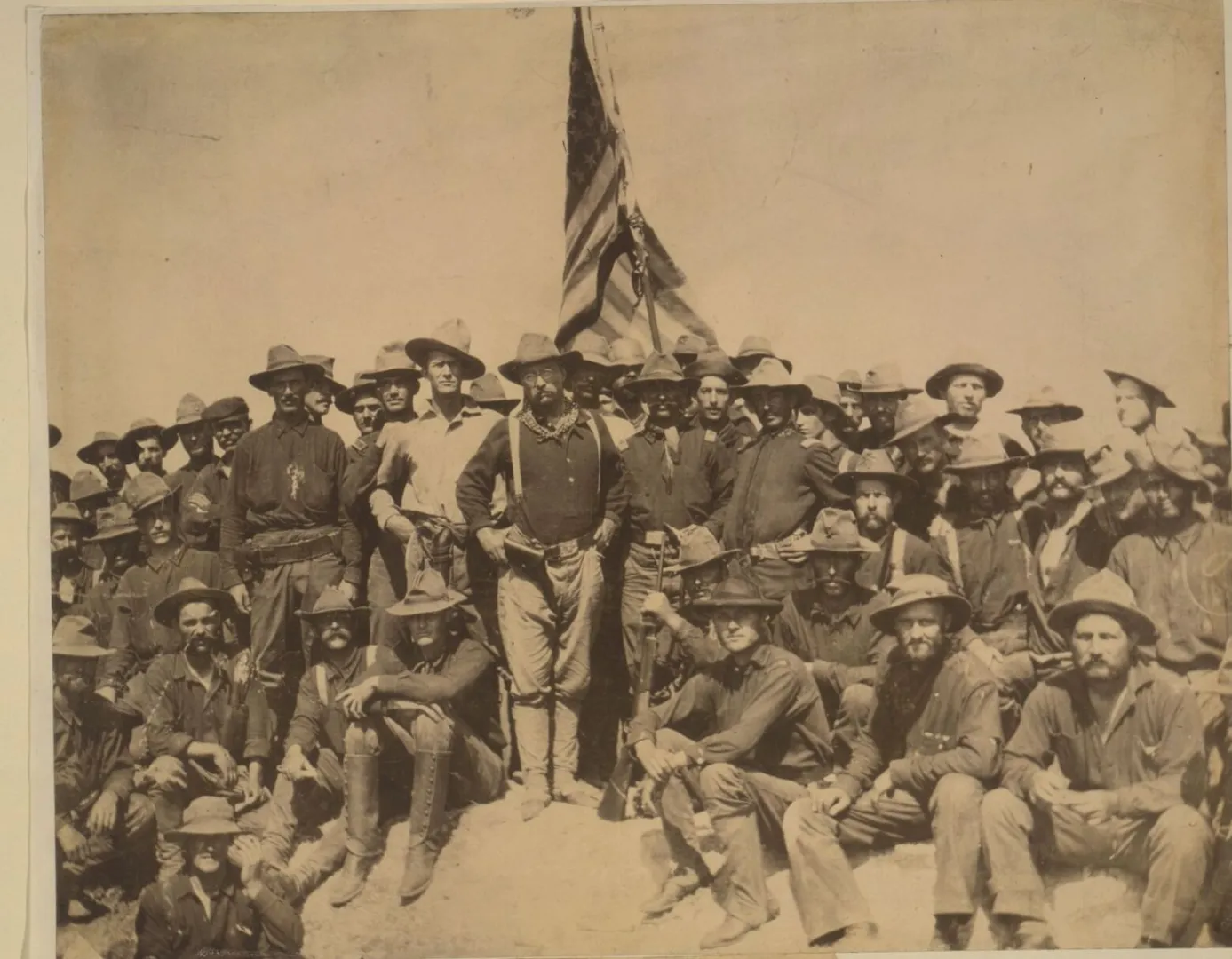 Colonel Roosevelt standing with the Rough Riders and an American flag atop the captured hill