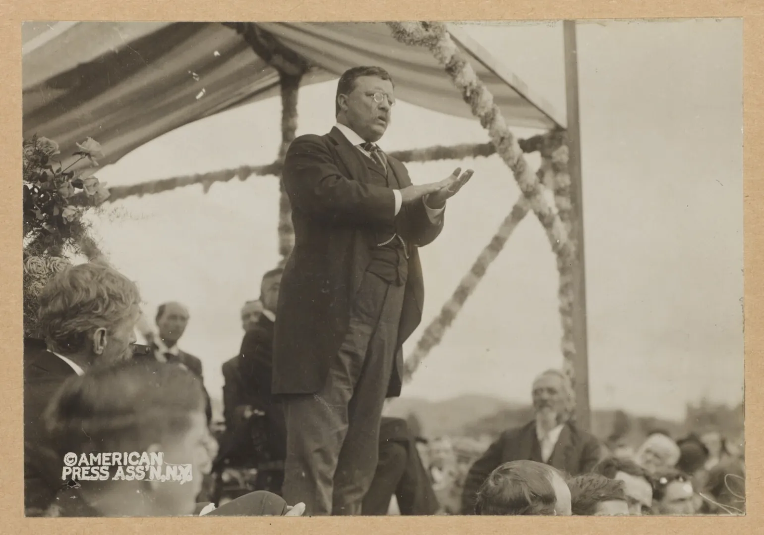 Theodore Roosevelt stands beneath a canopy, gesturing as he addresses a seated audience during his 1910 speaking tour