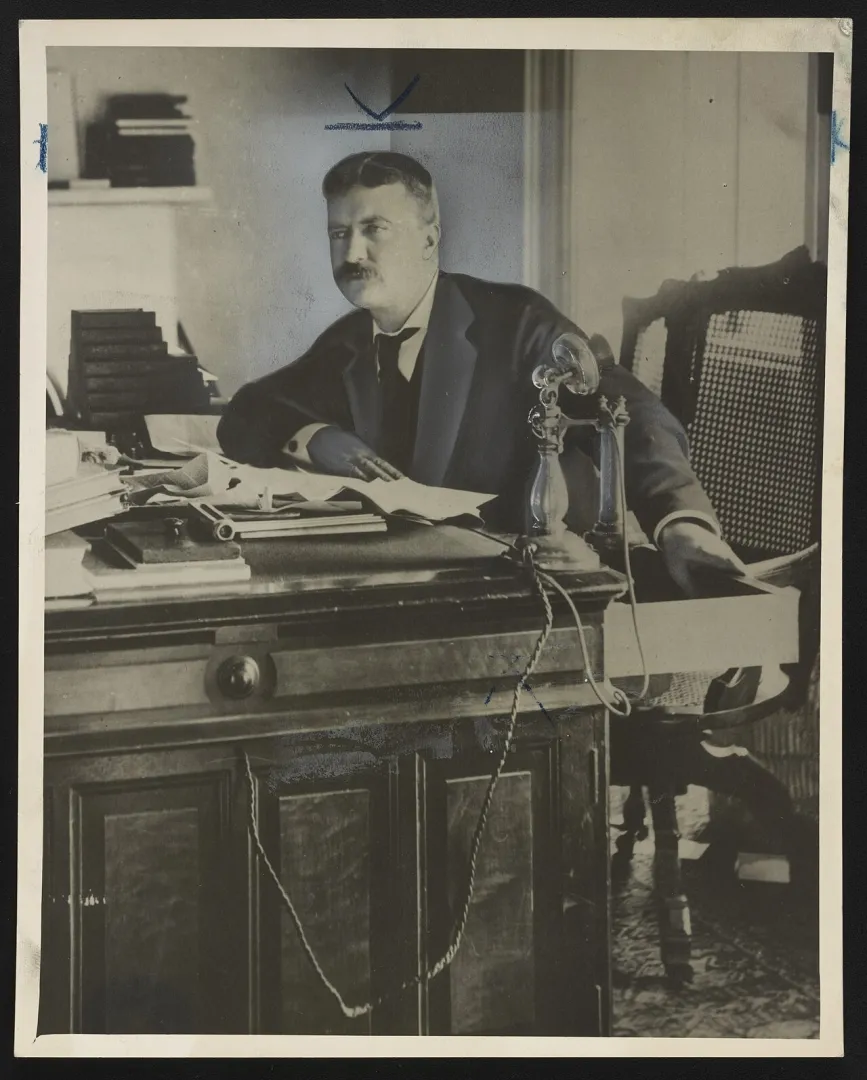 Theodore Roosevelt seated at a desk with papers and a candlestick telephone