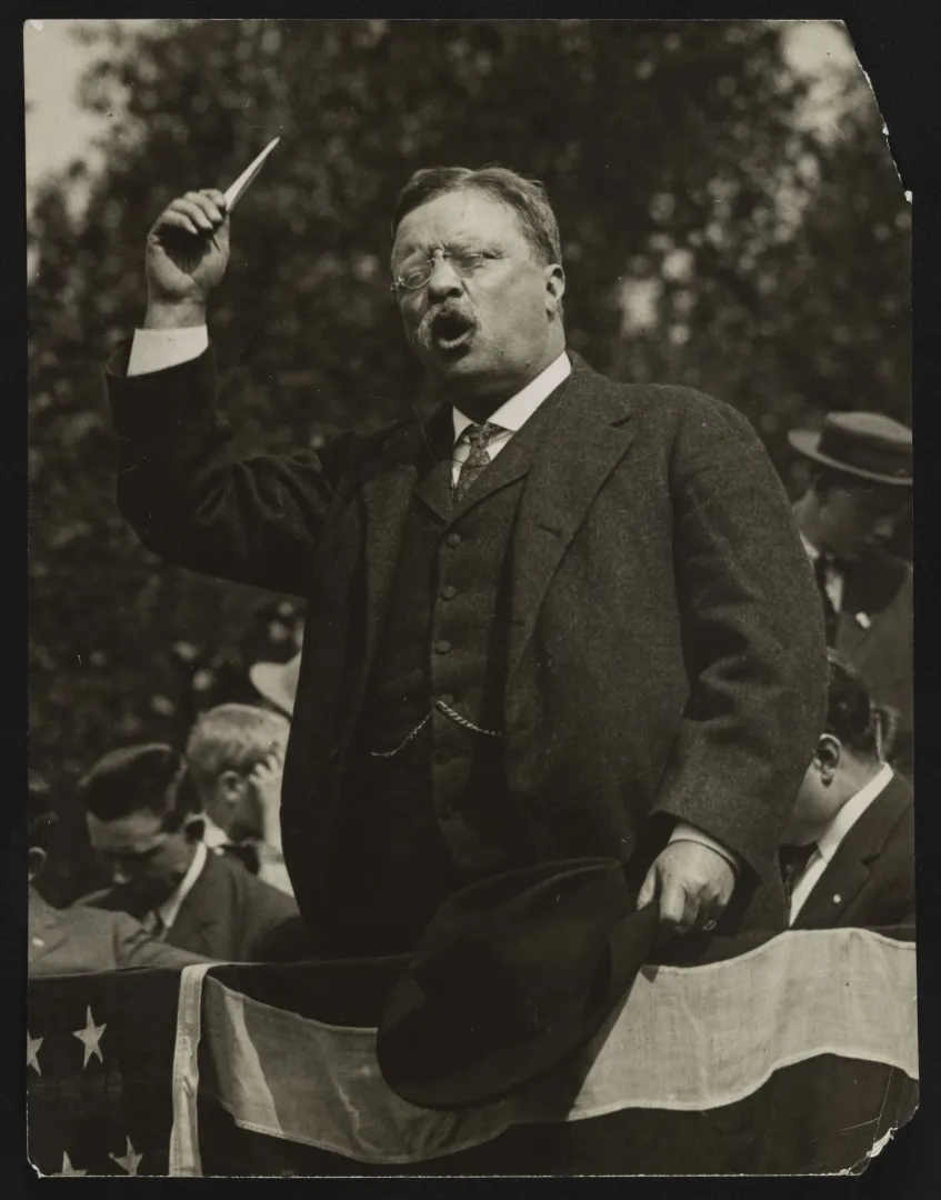 Theodore Roosevelt gesturing with a raised arm while speaking from a flag-draped platform