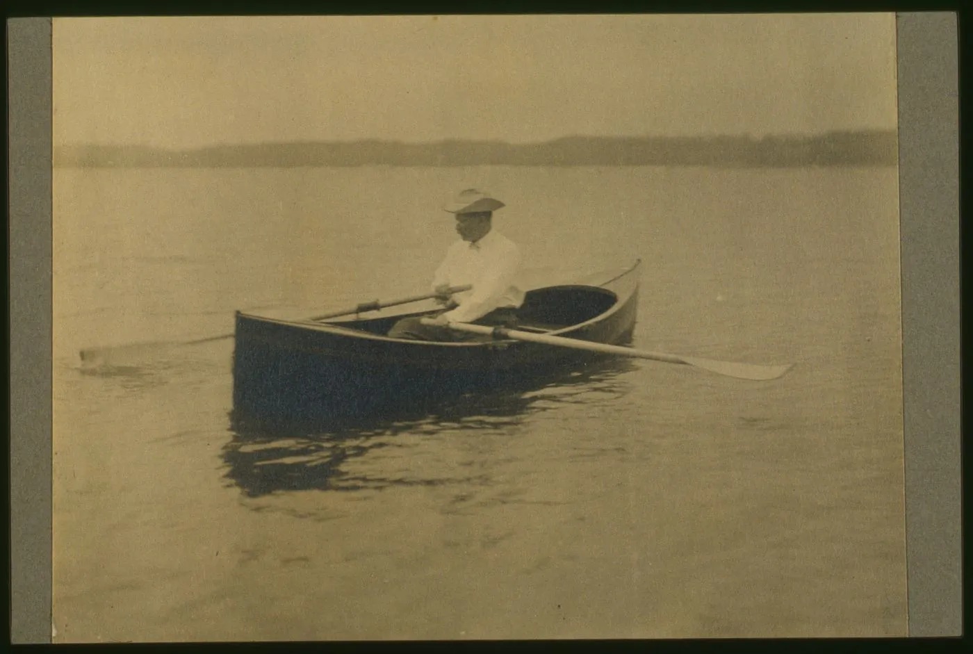 Theodore Roosevelt seated in a rowboat, pulling the oars across calm open water.