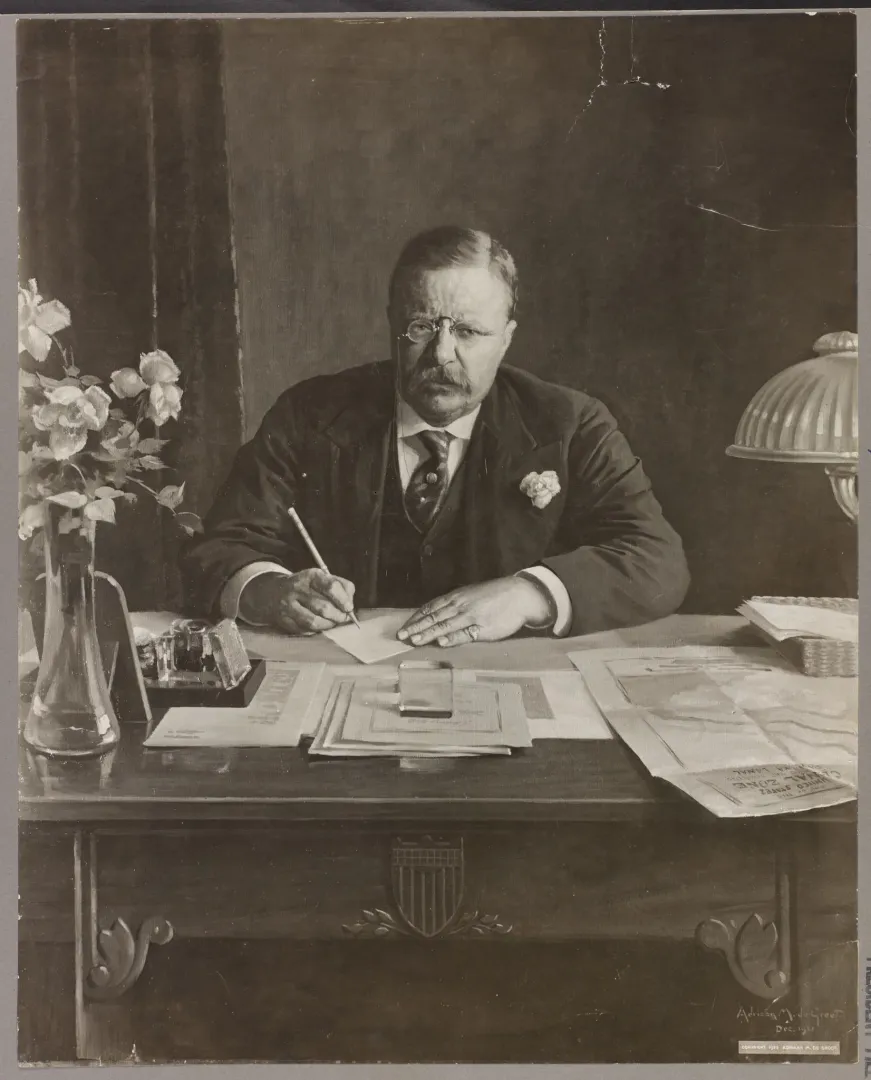 Theodore Roosevelt seated at his desk writing with a pen