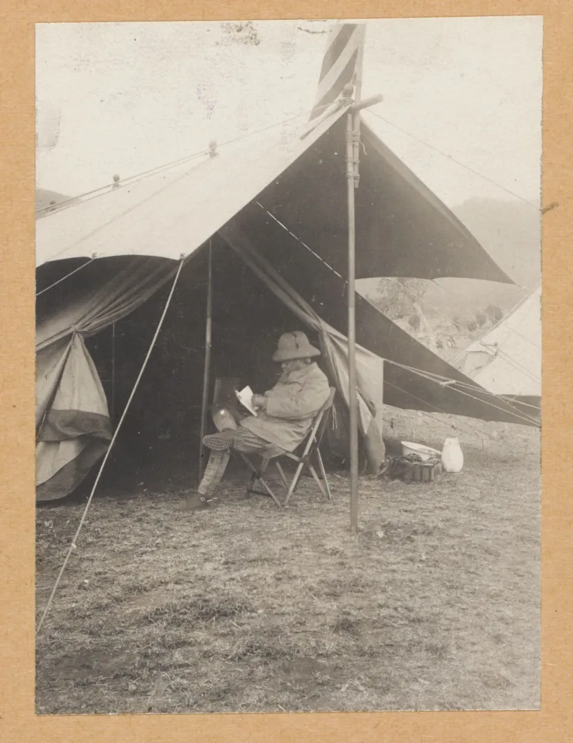 Theodore Roosevelt seated in a camp chair reading at the entrance of his tent