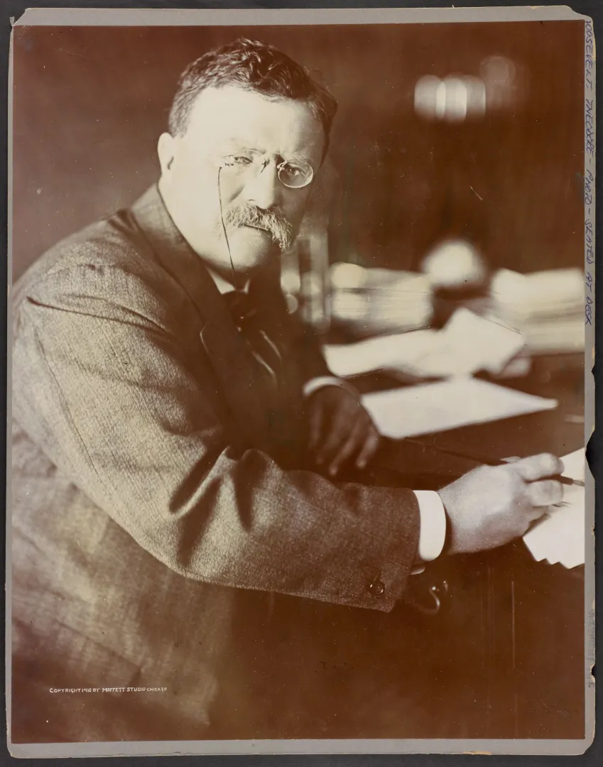Theodore Roosevelt seated at a desk holding a pen over papers
