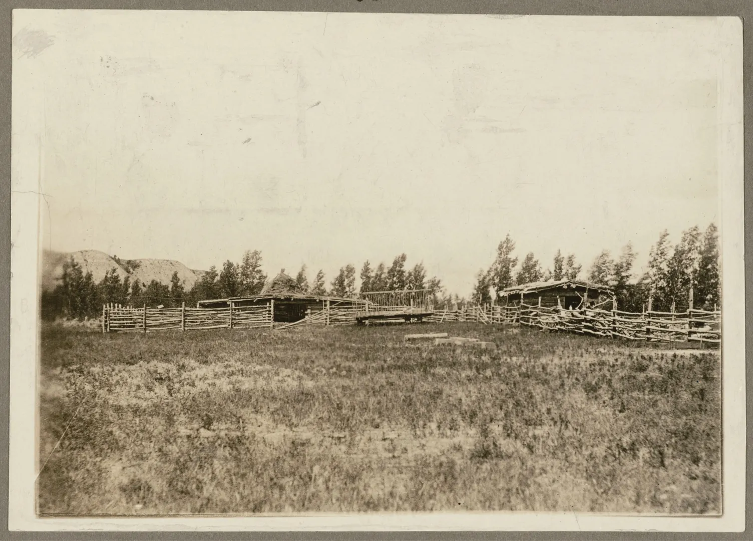 Historic photograph of the corrals and low ranch buildings at the Elkhorn Ranch, standing on open bottomland with a line of trees and bluffs behind